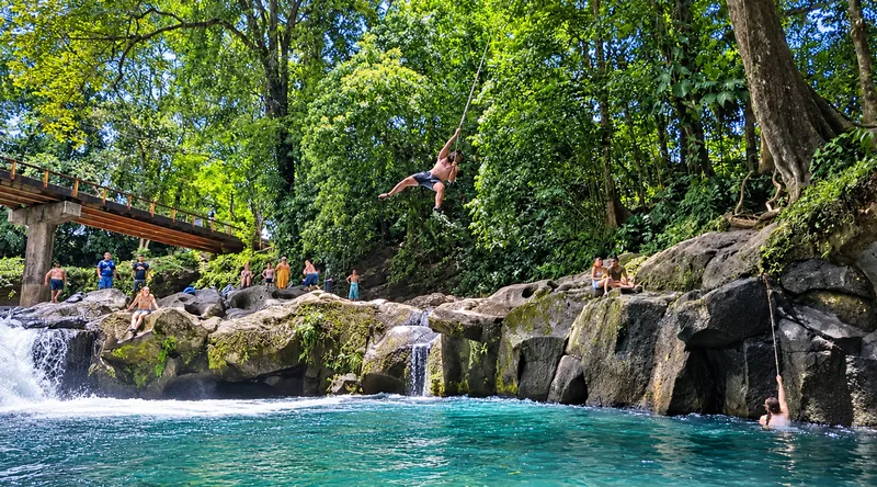 The whole spot: two ropes, the rocks people jump from, and the road bridge above. On a warm afternoon it is busy.